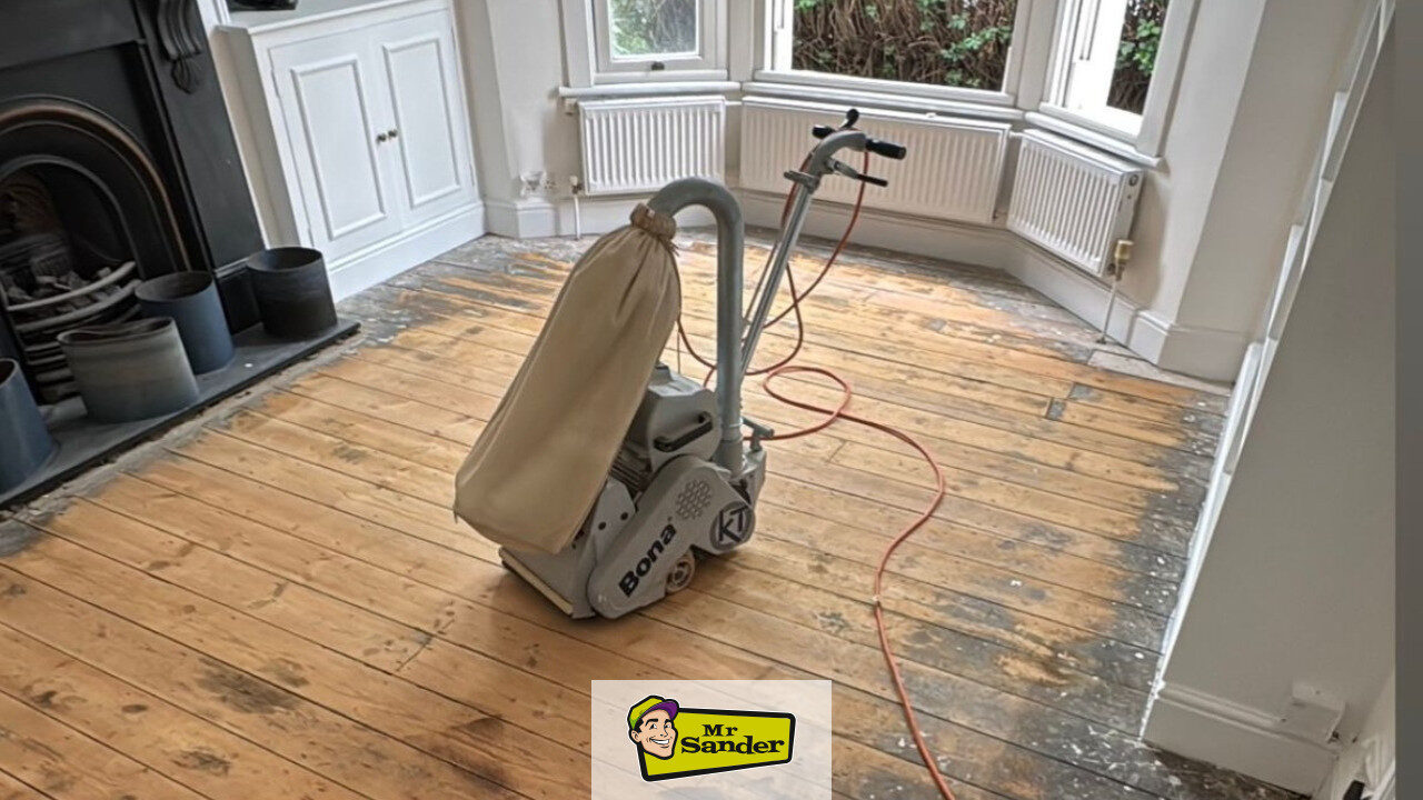 DIY floorboard sanding in a bay-window living room—pine boards mid-strip with darker edges, Bona drum sander with dust bag on site, white walls and radiator—Mr Sander®.