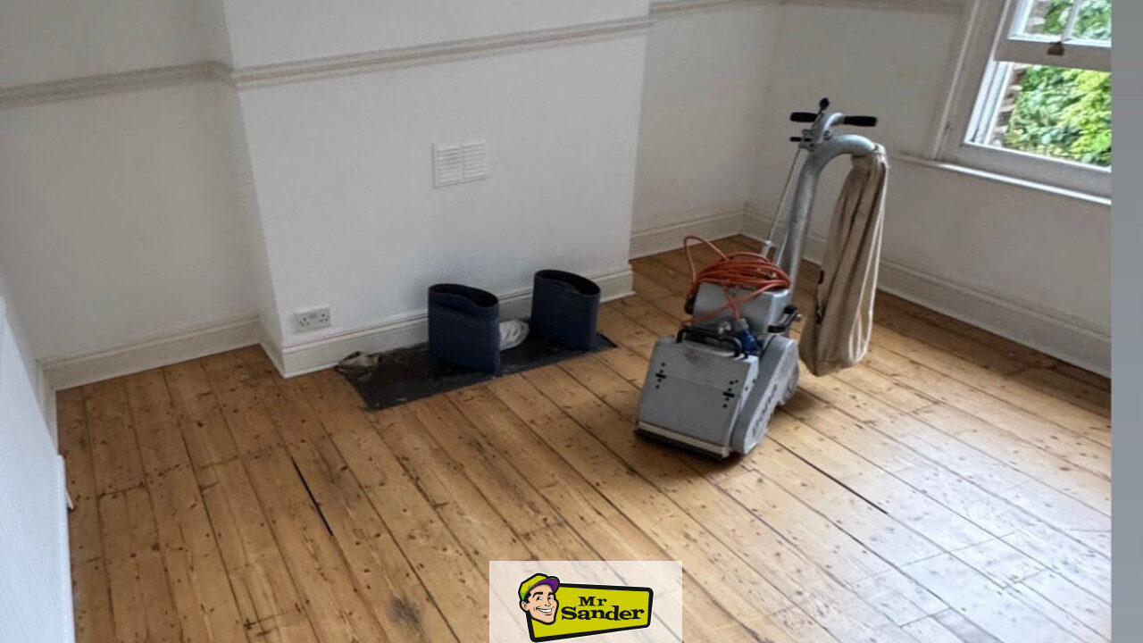 DIY floorboard sanding in a light living room—freshly sanded pine boards, drum/belt sander with dust bag by the bay window, white walls and small hearth—Mr Sander®.