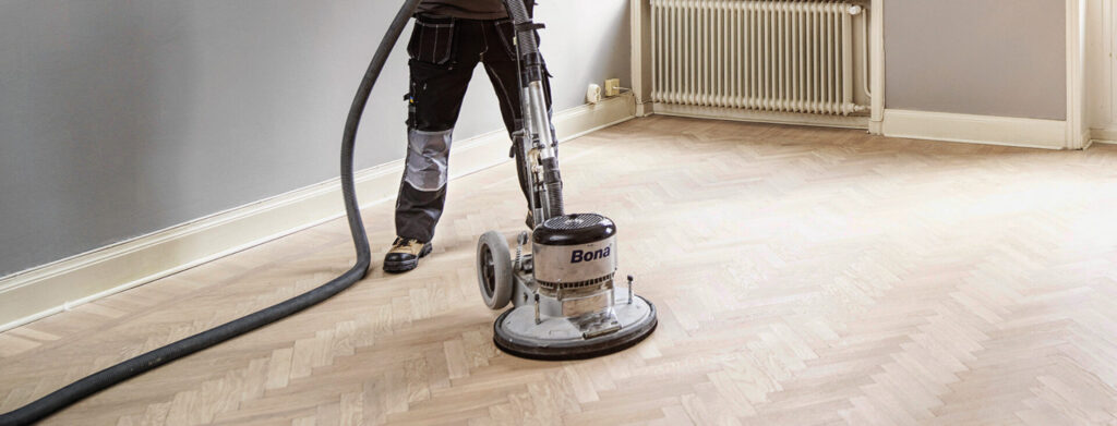 Technician operating a Bona rotary sander with dust-extraction hose on a light herringbone parquet floor.