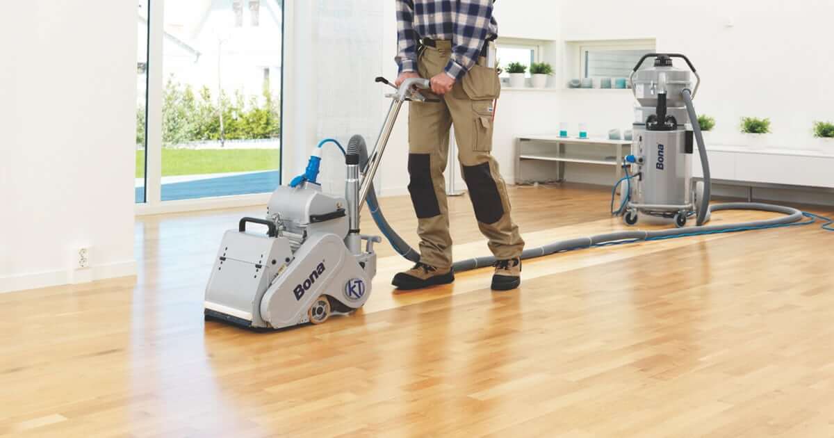 A man from Mr Sander® is sanding a floor in a living room in the UK using professional machines, including a Bona Belt AMO110000.3 with a 2.2 kW motor, 230 V, 50 Hz, and a drum width of 200 & 250 mm. The room has green hanging plants, blue columns, and a bar with green tiles. Various tools and supplies are visible, indicating ongoing work. Dust containment is managed by a Bona DCS 70 with a 70 L capacity, HEPA fine filter, and 235 m³/hour airflow.