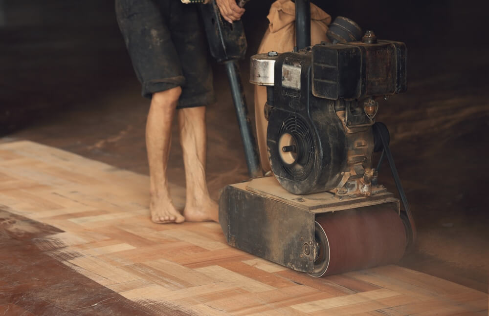A person using a large floor sanding machine to sand and refinish a wooden parquet floor. The individual is wearing rugged clothes and is barefoot, with dust surrounding the area.