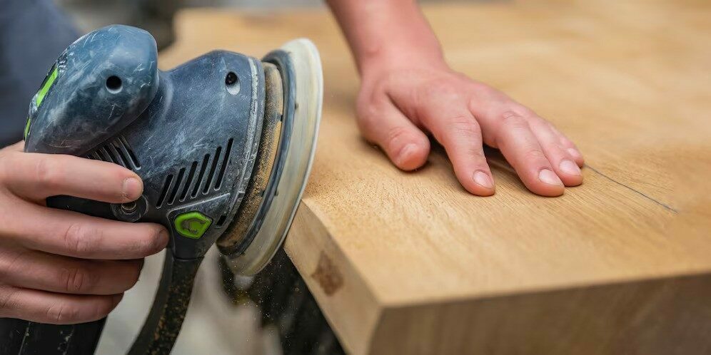 Close-up of a hand guiding an orbital sander over a wooden surface.