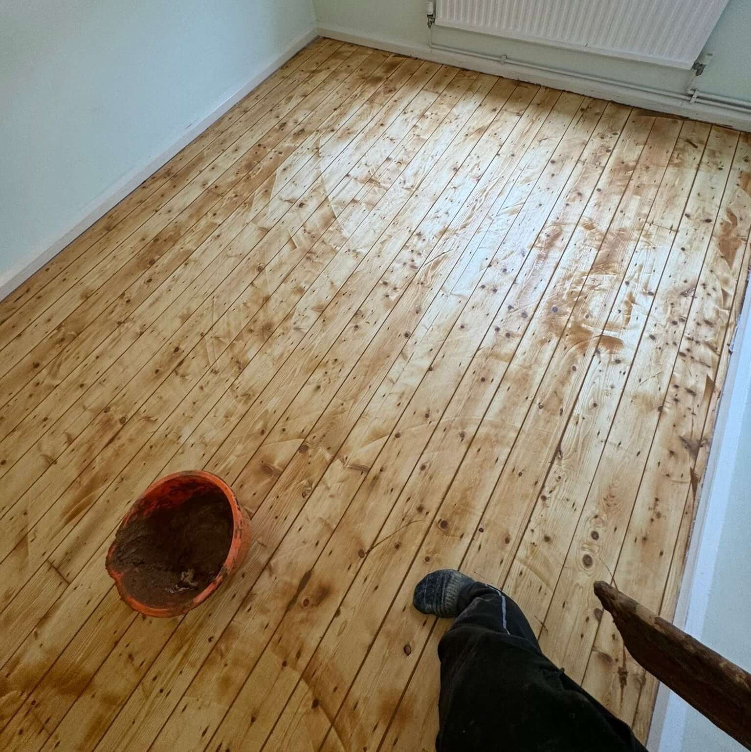 A pine wood floor in the process of being refinished, with gaps and cracks being filled with a wood filler. An orange bucket filled with wood filler sits on the floor, and a person’s foot is visible in the corner. The floor sanding and refinishing are being handled by Mr Sander®.