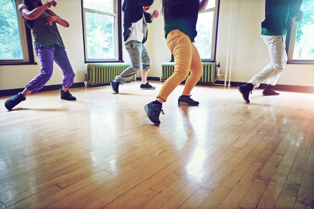 Group of dancers rehearsing hip‑hop moves on a pale maple sprung floor in a light‑filled studio, showcasing professional floor installation that balances resilience and glide.