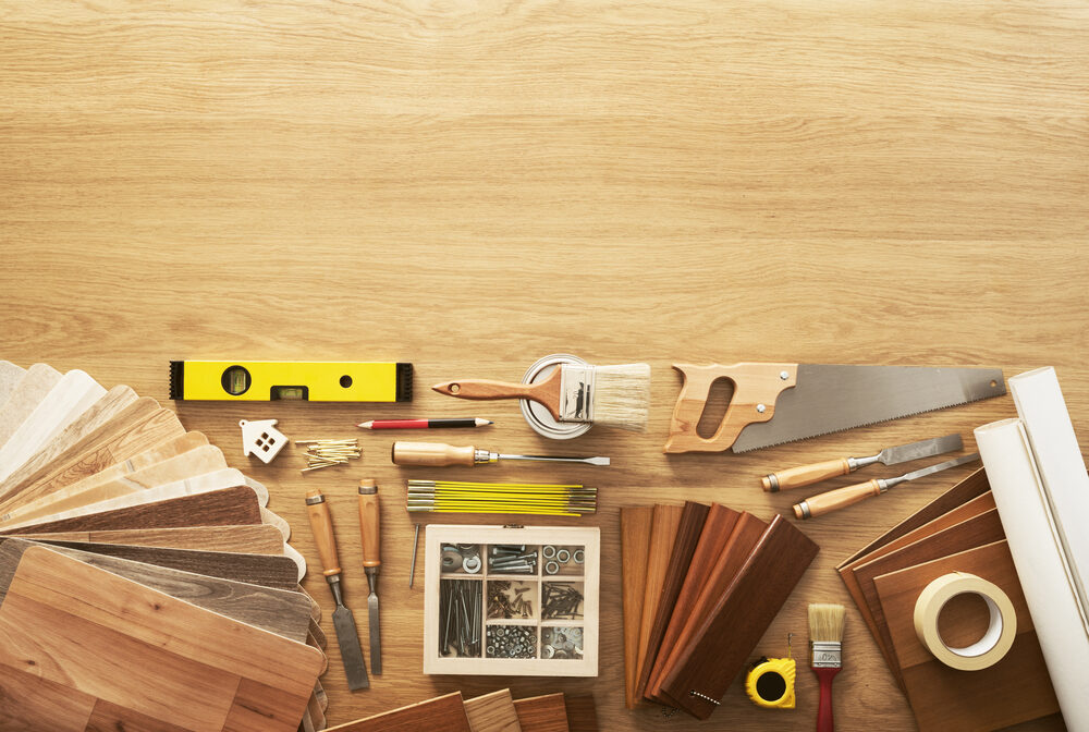 Flat-lay view of floor restoration equipment: spirit level, hand saw, chisels, paintbrush, nails, screws, measuring tape, masking tape and assorted hardwood flooring samples arranged on an oak work surface