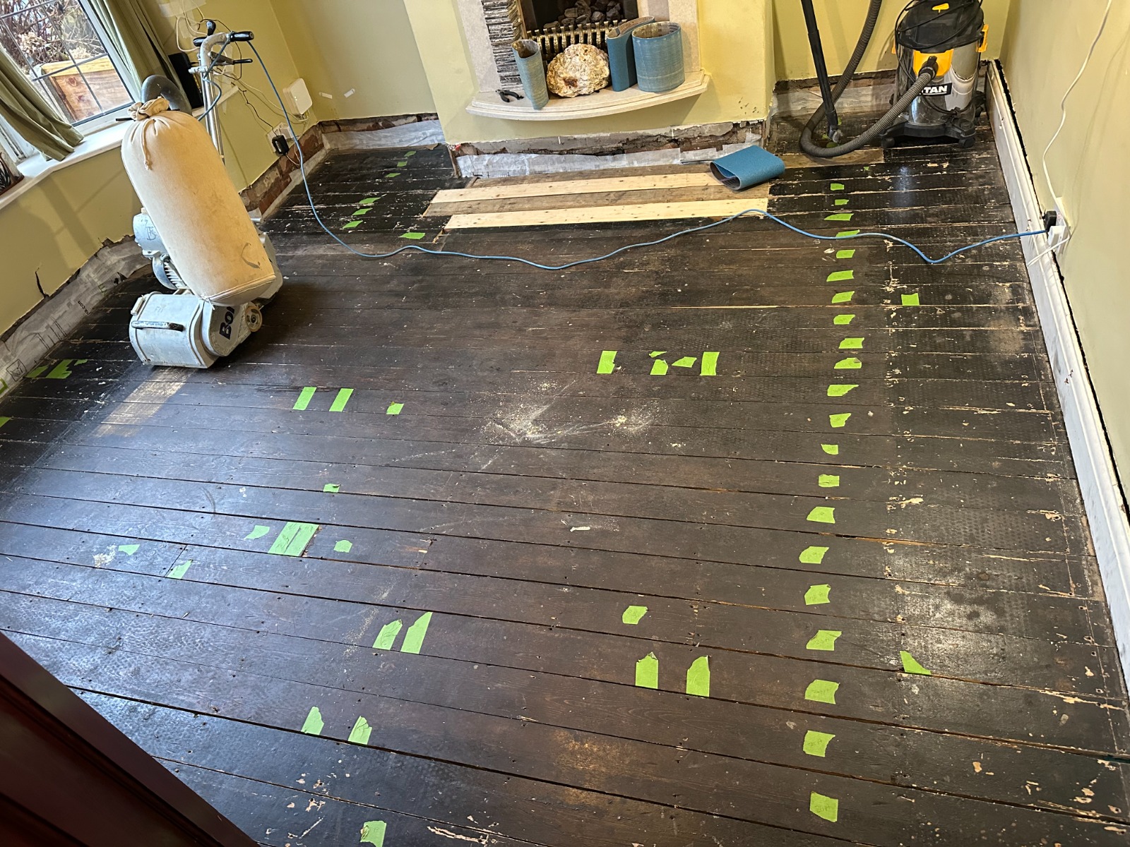 Floor Sanding in London on dark-stained solid pine floorboards, showing a living room prepared for restoration with a drum sander, vacuum and marked repair areas.