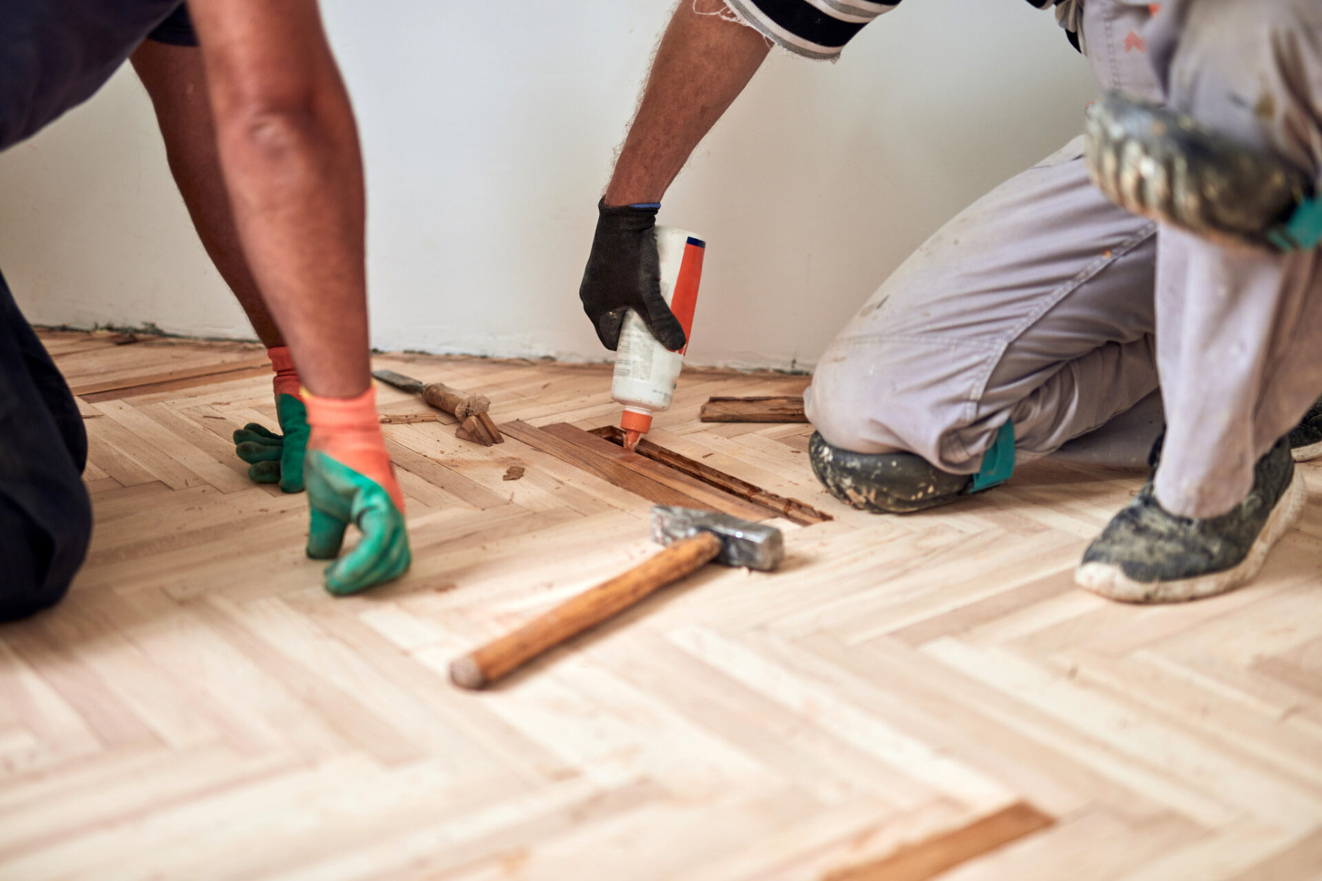 Floor Sanding in London with repairs to a light oak herringbone parquet floor, as technicians replace damaged parquet blocks using wood glue and tools.