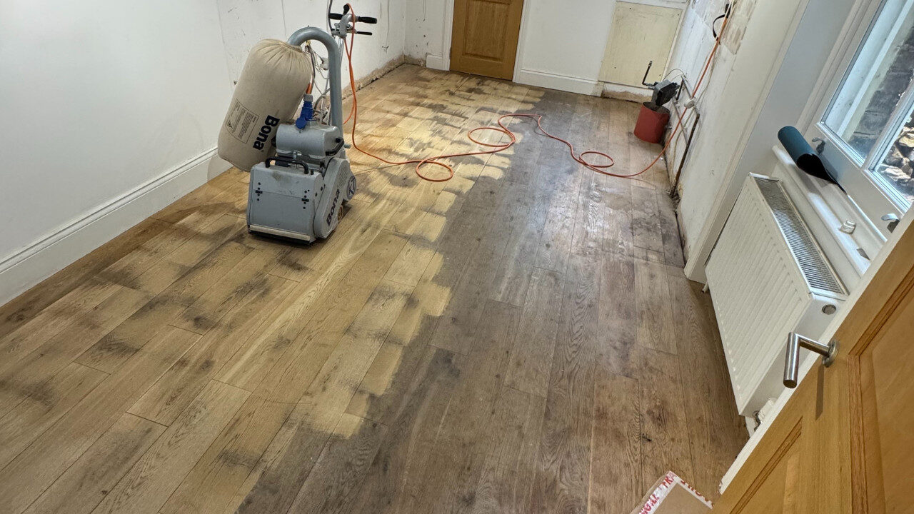 Oak plank kitchen floor mid-sanding with a Bona belt sander and dust bag, showing cleaned passes beside worn, darker boards and trailing power lead.