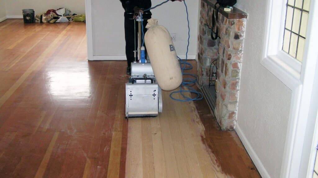 A professional from Mr Sander® is sanding a solid oak hardwood in a living room in, UK using a Bona Belt sander (AMO110000.3). The machine features a 2.2 kW motor, 230 V voltage, 50 Hz frequency, and a sanding belt size of 200 x 750 mm or 250 x 750 mm. The total weight is 74 kg.