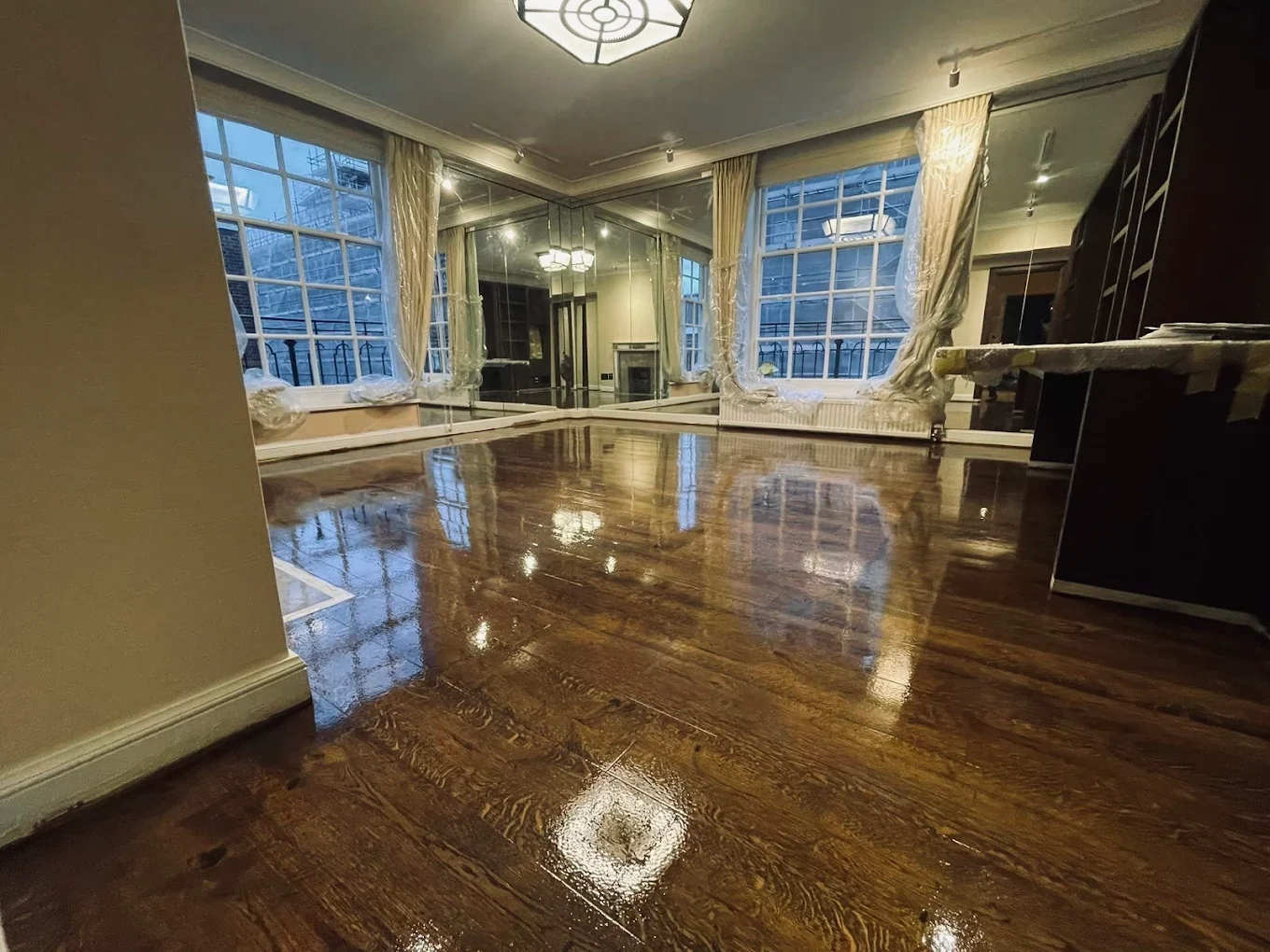 Luxury living room with large windows and mirrored wall showing oak floorboards professionally sanded, stained and finished to a high-gloss sheen, reflecting light across the room.
