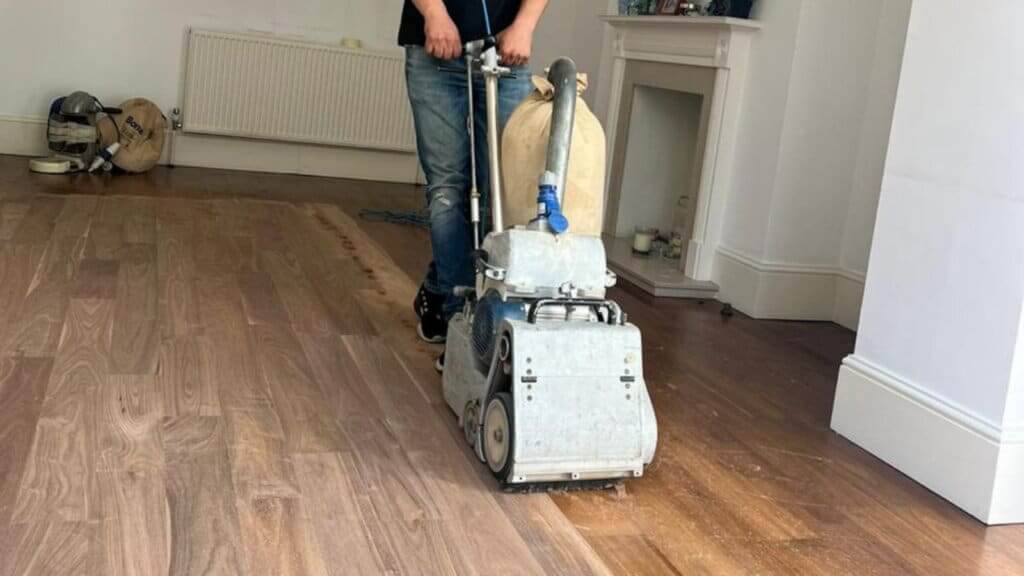 A professional from Mr Sander® is sanding a solid oak hardwood in a living room in Tingewick, UK using a Bona Belt sander (AMO110000.3). The machine features a 2.2 kW motor, 230 V voltage, 50 Hz frequency, and a sanding belt size of 200 x 750 mm or 250 x 750 mm. The total weight is 74 kg.