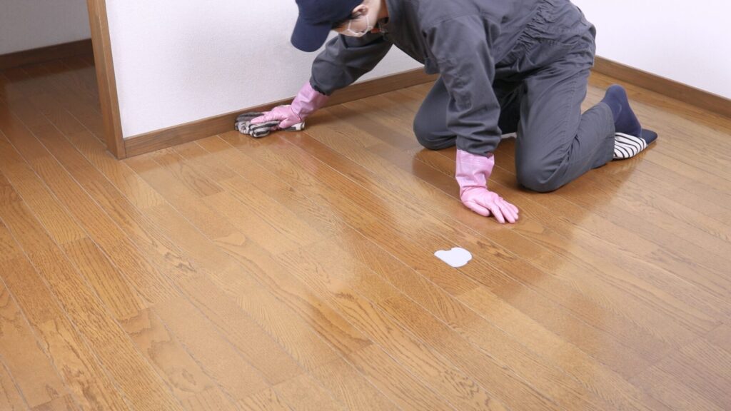Technician in coveralls and pink gloves hand-buffing solid oak strip flooring along the skirting board, final step to restore pet-stained floors.