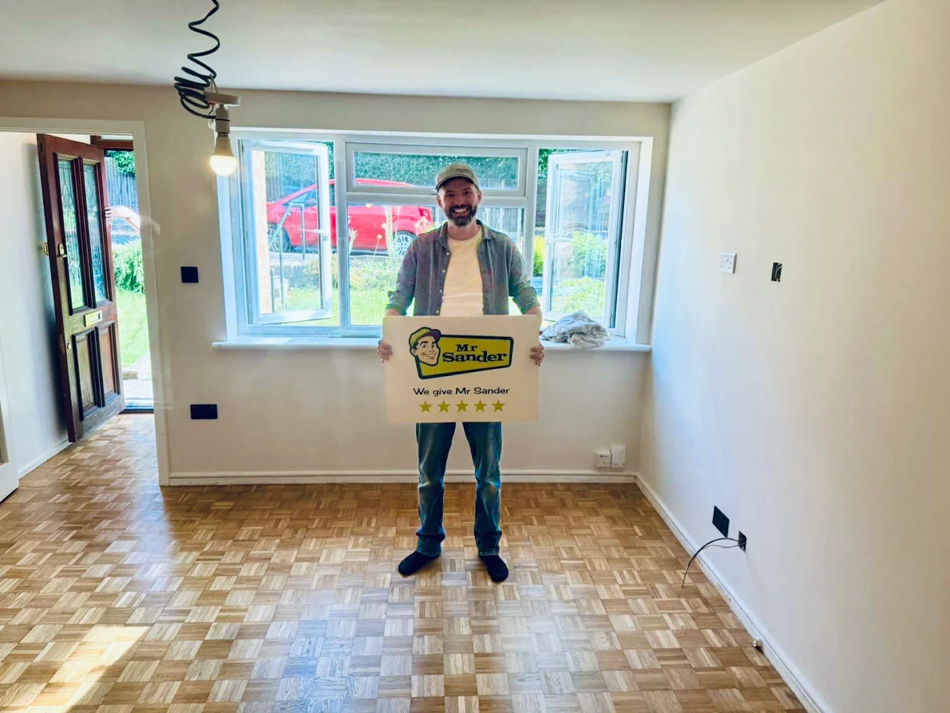  Smiling homeowner stands on a newly refinished mosaic parquet floor, holding a yellow Mr Sander sign that rates the service five stars.