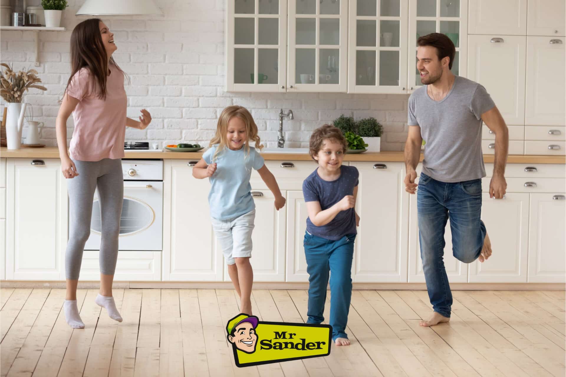 A family of four, including two children and two adults, joyfully dance in a bright kitchen with light-colored wooden floors. The kitchen features white cabinets, a wooden countertop, and a white brick backsplash. The floor is freshly sanded and refinished, enhancing the natural wood grain. A logo for Mr Sander® is visible in the bottom corner.