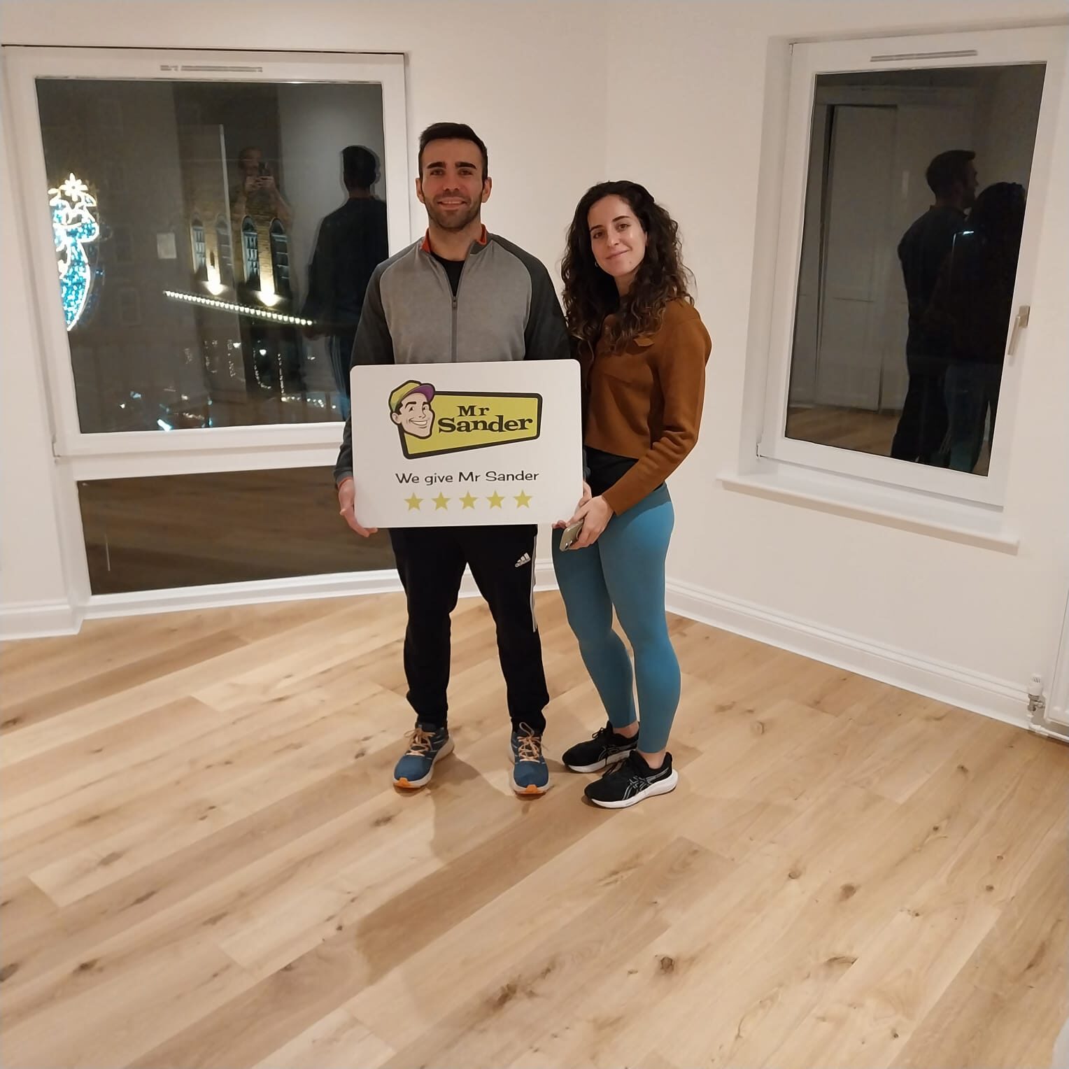 Happy customers holding a Mr Sander review sign while standing on a newly refinished oak engineered wood floor, praising workmanship on Sports Hall Lines and wood finishing.