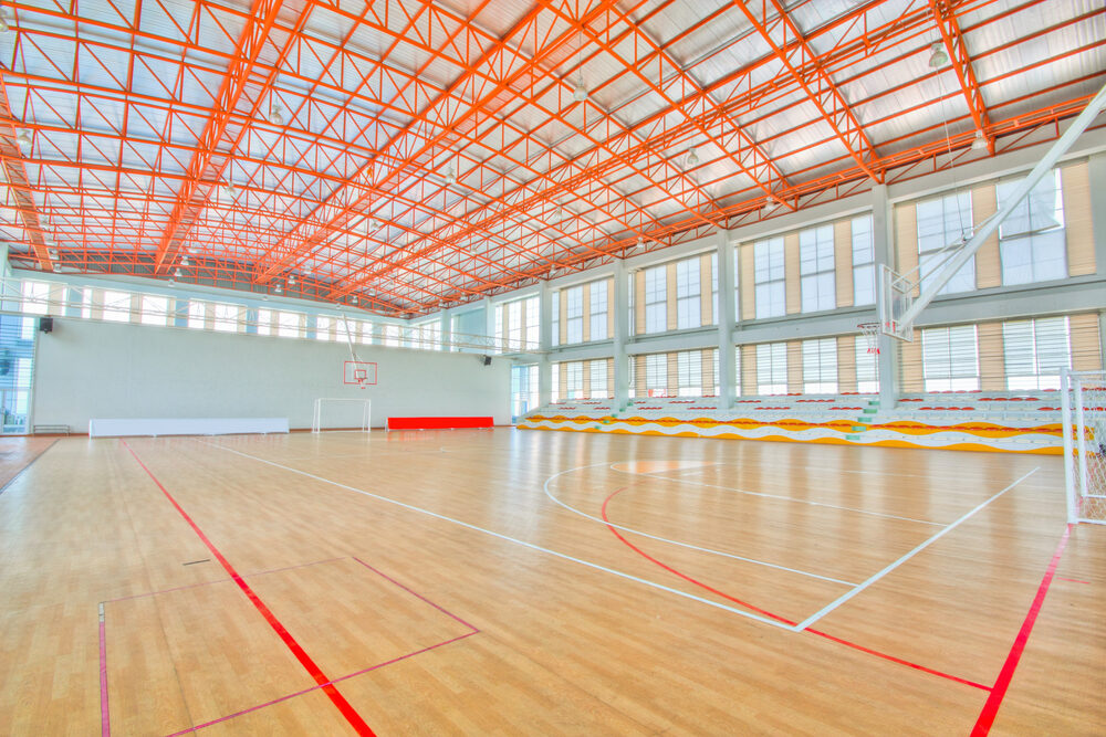Bright indoor sports hall with fitted hardwood floor, red and white basketball lines, and an orange steel roof with tiered seating.