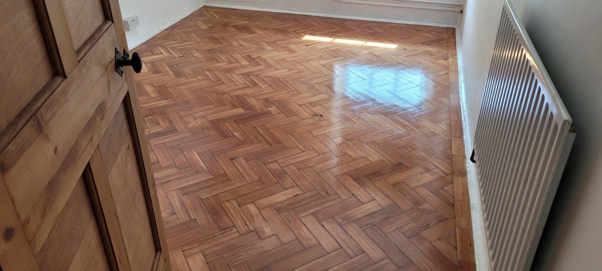  Freshly sanded and lacquered herringbone parquet floor in a small bedroom, viewed from an open wooden door with a radiator along the right‑hand wall.