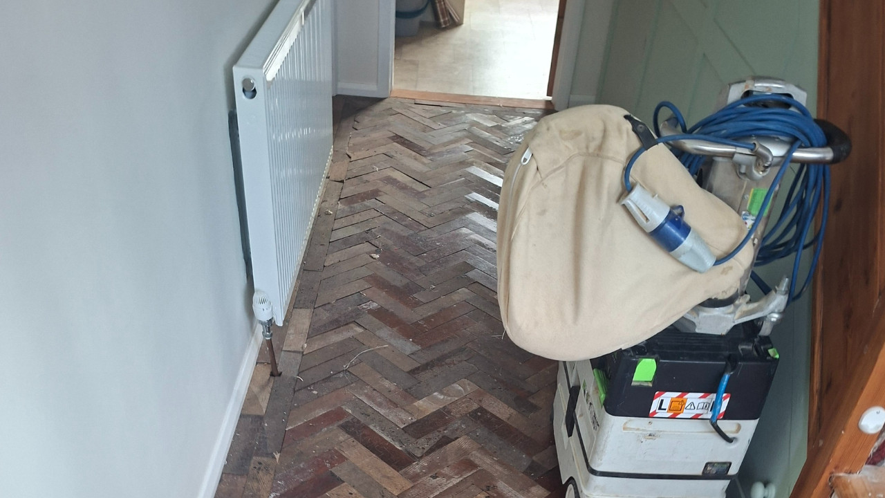 A worn oak herringbone parquet floor in a hallway during the initial stages of a professional herringbone sanding project.