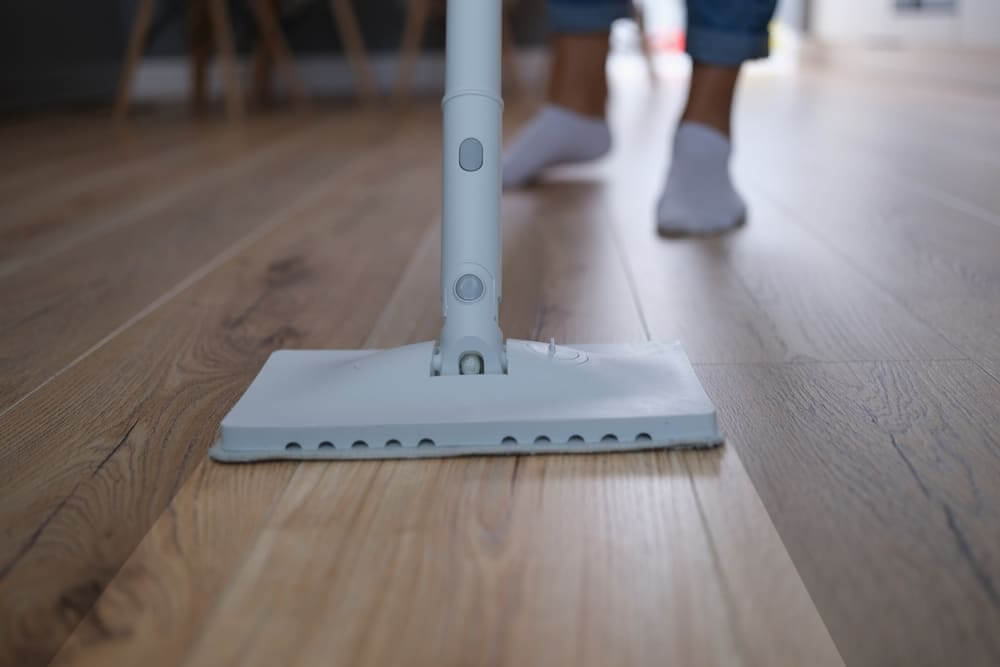 A close-up view of a person using a mop to clean a shiny, freshly refinished wooden floor. The mop head glides over the floor, highlighting the smooth, polished surface.