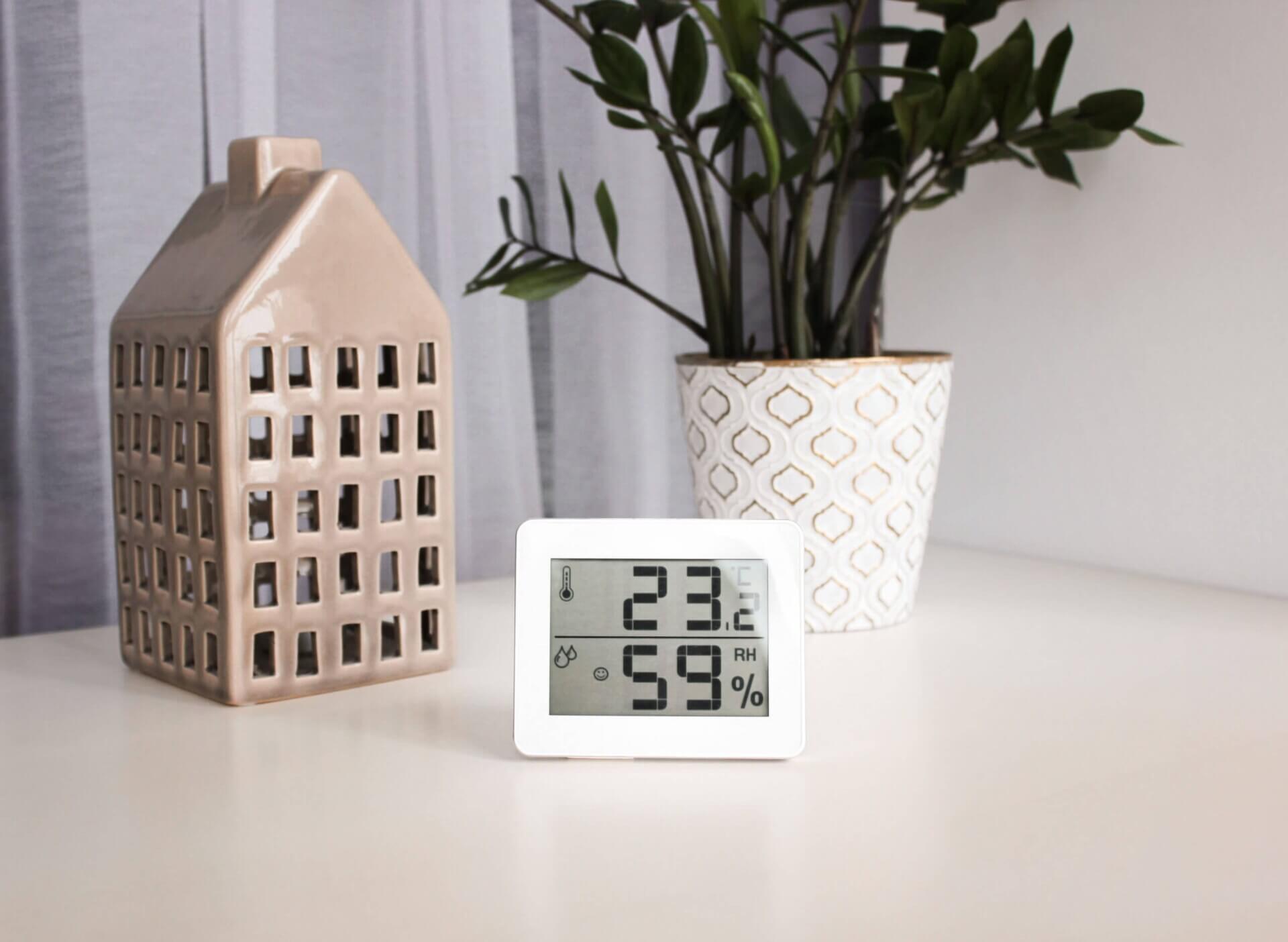 A digital hygrometer displaying temperature and humidity levels on a table with a decorative ceramic house and a potted plant in the background.