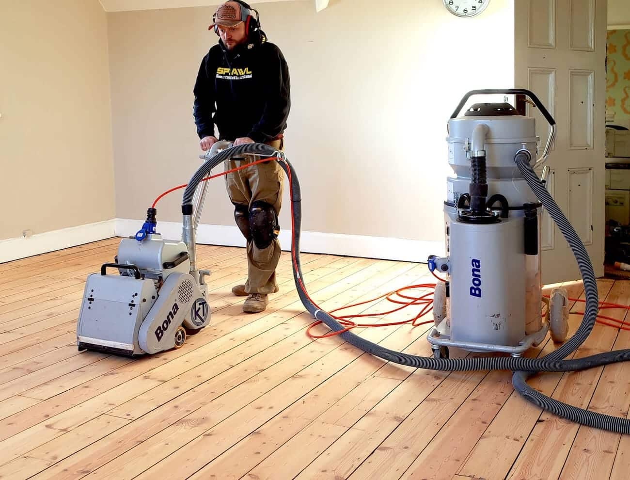 A professional from Mr Sander® sanding a pine floor in a living room in Surrey, UK using the Bona Belt sander and Bona DCS 70 dust containment system. The Bona Belt features a 2.2 kW motor, 230 V voltage, 250x750 mm belt size, and weighs 74 kg. The Bona DCS 70 has a 70 L dust capacity and HEPA filter.
