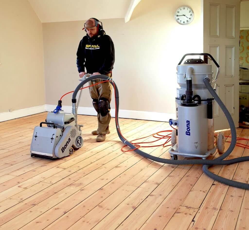 A Professional from Mr Sander® sanding a pine floor in a living room. The equipment includes a Bona Belt sander (AMO110000.3) with 2.2 kW motor power, 230V voltage, 50 Hz frequency, 250x750 mm size, and 74 kg weight. Additionally, a Bona DCS 70 (AMO530005) dust containment system is used, featuring a 230V motor, 1400W power, 70L dust holding capacity, HEPA class H 13 filter, 7.5m hose length, and 43 kg weight.
