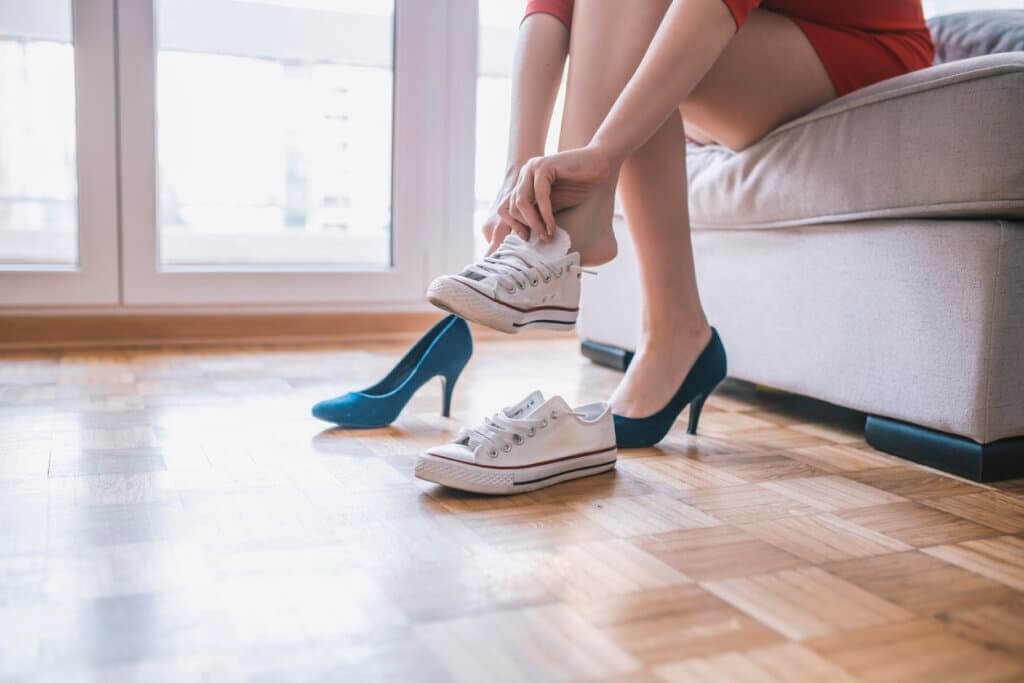 A woman sits on a sofa in a brightly lit room with large windows, changing from white sneakers to blue high heels. The parquet wooden floor beneath her is beautifully finished, showcasing the high-quality floor sanding and refinishing services provided by Mr Sander®.