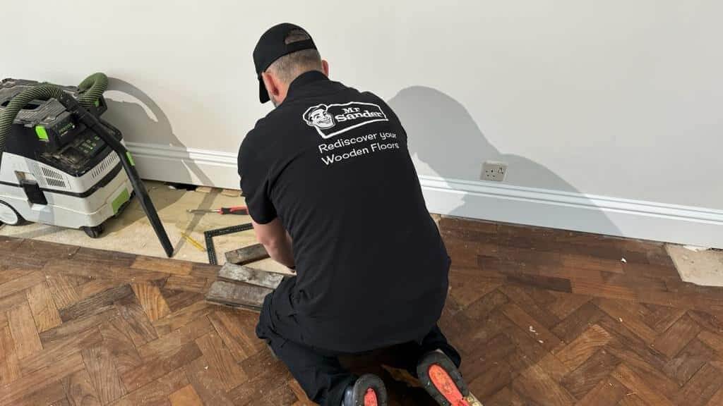 A Mr Sander® professional in a black uniform is seen from behind, diligently working on parquet flooring restoration. The scene includes various tools and a vacuum system, emphasizing the detailed process involved in restoring the elegance of parquet floors. This image is part of the blog post titled "Reclaim Your Home's Elegance: The Untold Secrets to Flawless Parquet Flooring Restoration," showcasing the expertise of Mr Sander® in parquet flooring restoration.