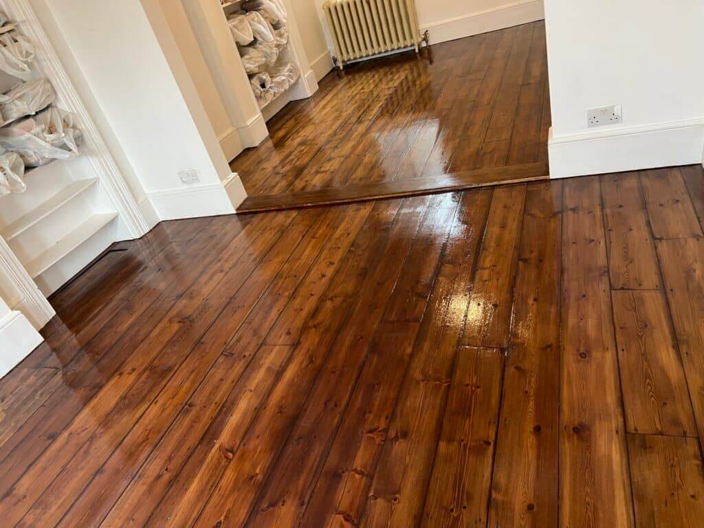 A room with dark wooden floors that have been freshly sealed, giving them a rich, glossy finish. The floor reflects light, highlighting the smooth and polished surface. The room features white walls, built-in shelving, and a radiator in the background. The area transitions seamlessly from one room to another, both showcasing the same high-quality finish.