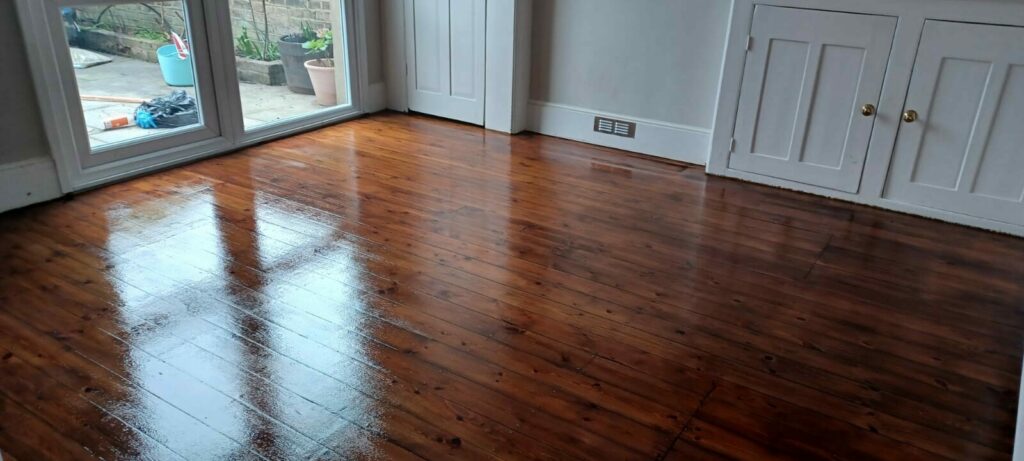 Reflective wooden floor in a room with double doors opening to an outdoor area, with cleaning supplies visible outside.
