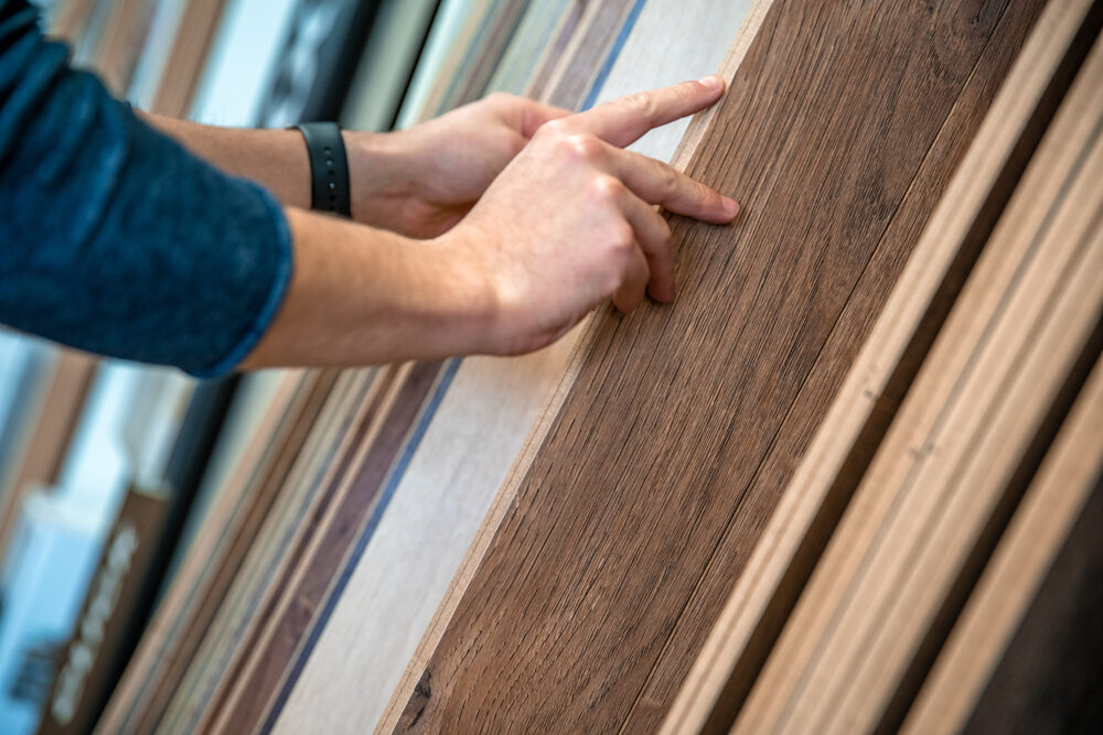Close‑up of a person’s hands feeling the grain of a dark‑oak floorboard sample in a showroom display, comparing finishes and colour tones.