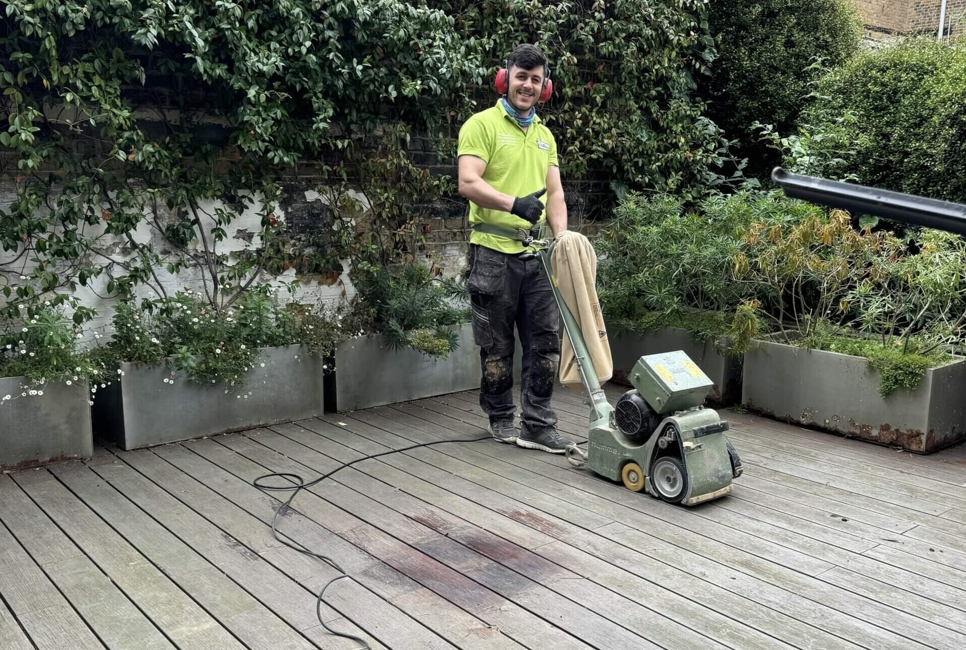 Joyful Decks sanding in progress: technician operating a professional drum sander with dust bag on weathered smooth timber decking, preparing the boards for refinishing.