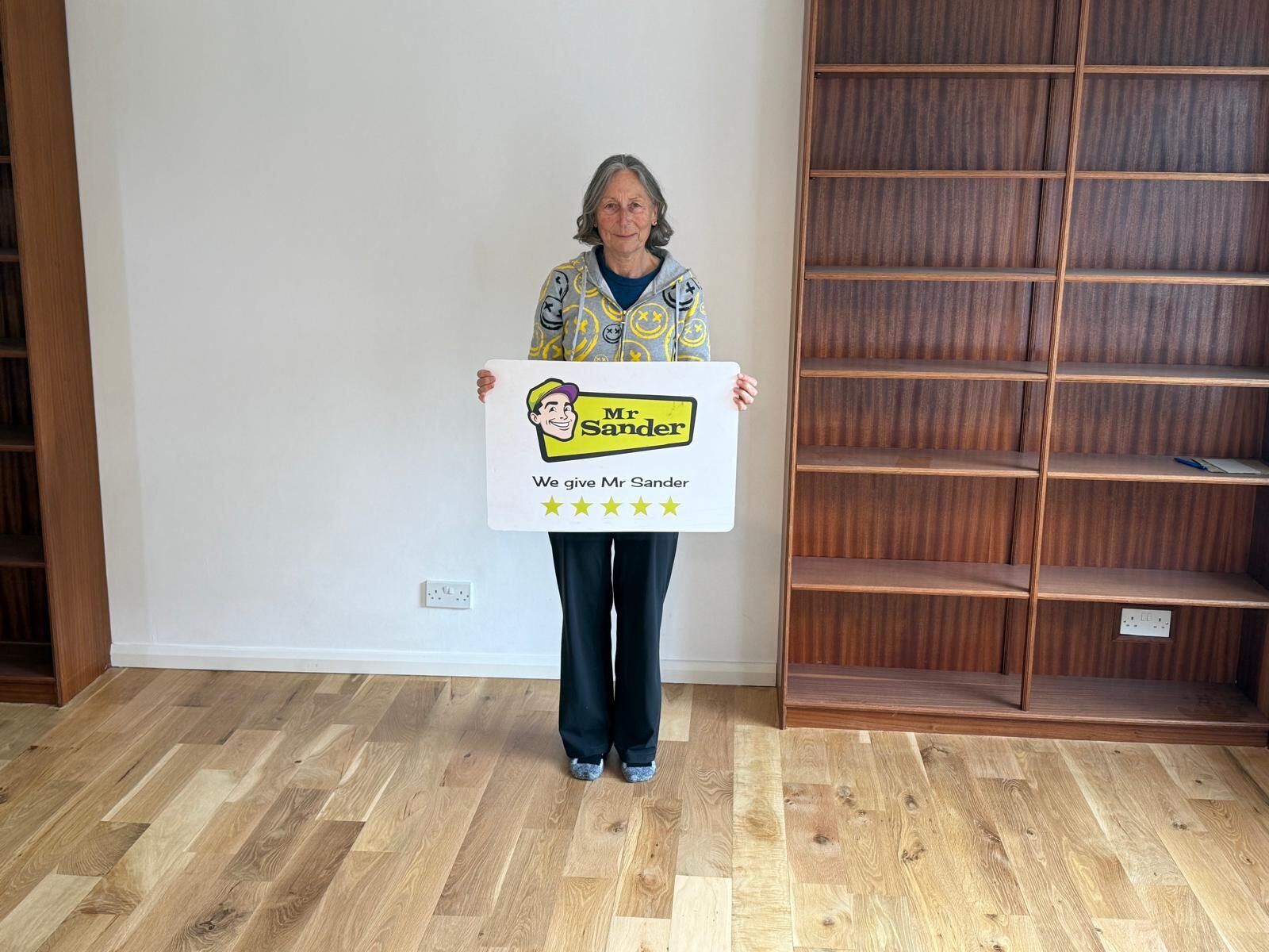 Customer holding a Mr Sander® sign on a newly installed oak solid hardwood floor, showcasing premium Junckers Floor Fitting quality and finish.
