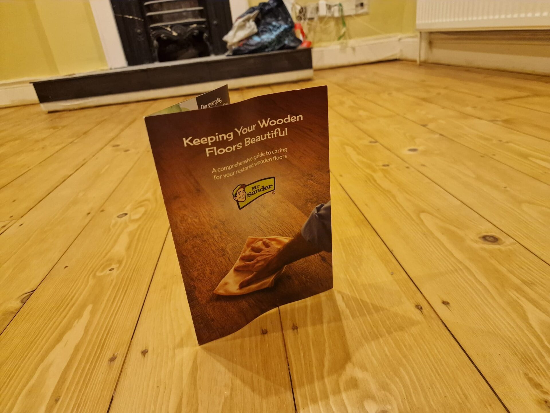 Mr Sander® “Keeping Your Wooden Floors Beautiful” care guide standing on a newly sanded and finished pine floor in a living room.