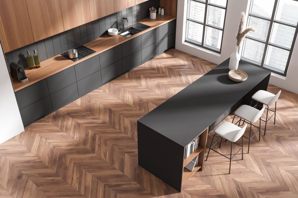A modern kitchen with a beautifully refinished chevron-pattern parquet wood floor, featuring a warm brown tone. The kitchen has sleek, dark cabinetry with wood countertops, a minimalist island with bar stools, and large windows allowing natural light to brighten the space.
