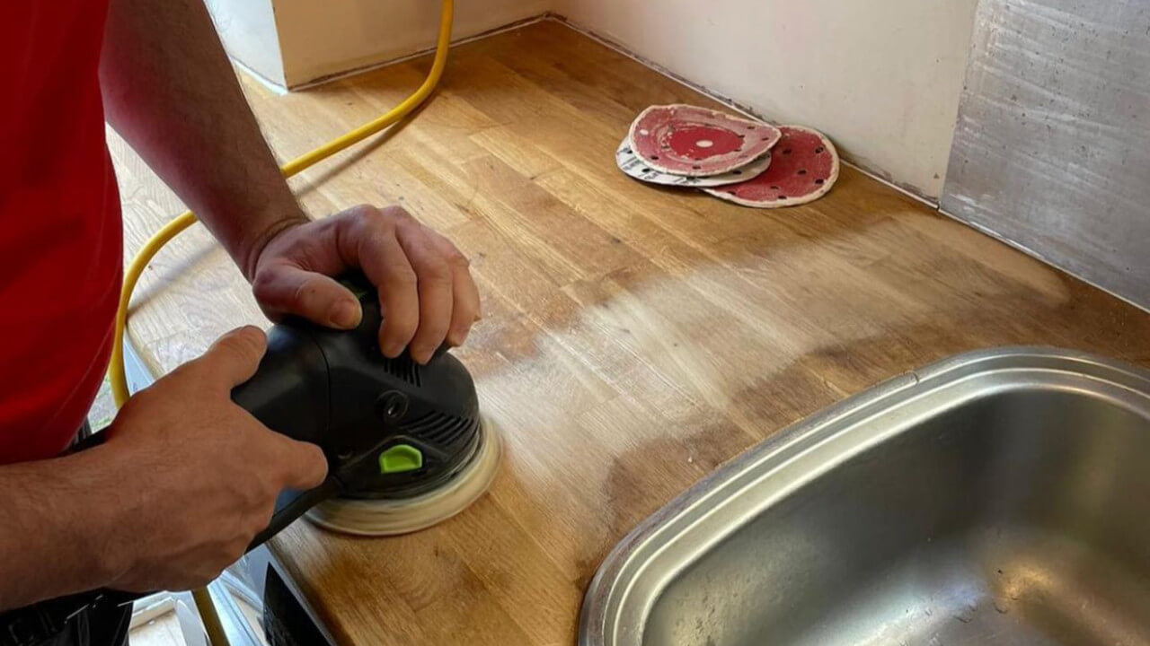 Teknisyen, altında meşe parke döşeme tahtaları bulunan, paslanmaz çelik bir lavabonun yanındaki meşe kasap bloklu tezgahın üzerinde elektrikli zımpara makinesiyle Mutfak Tezgahı Zımparalama işlemini gerçekleştiriyor.