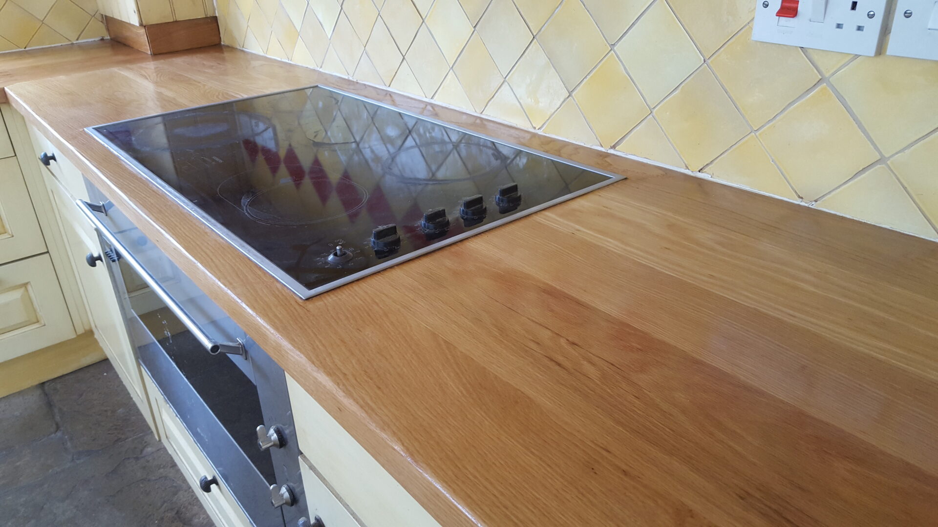 Kitchen Worktop Sanding results on a smooth solid oak hardwood worktop around an electric hob, next to a natural stone tile floor and tiled splashback.