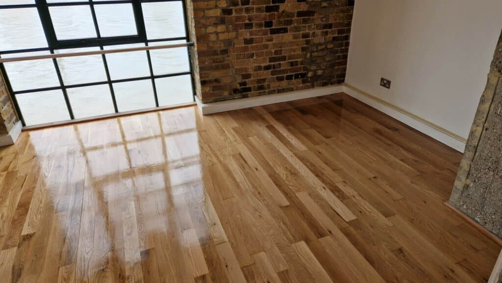 Newly refinished wooden floor in a room with brick walls and large windows, reflecting natural light.