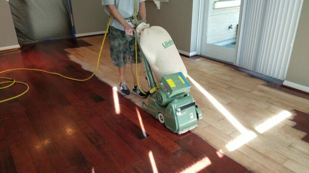 A man from Mr Sander® sands a floor in a living room in Tonbridge, UK, using a Lagler HUMMEL® sanding machine. The machine features a 230 V single-phase AC motor with 2.2 kW power, 2400 rpm drum speed, 200 mm drum width, 200 x 750 mm sanding belt, and weighs 79 kg. It includes a powerful LED lighting system and provides high powered aggressive sanding with low dust emissions.