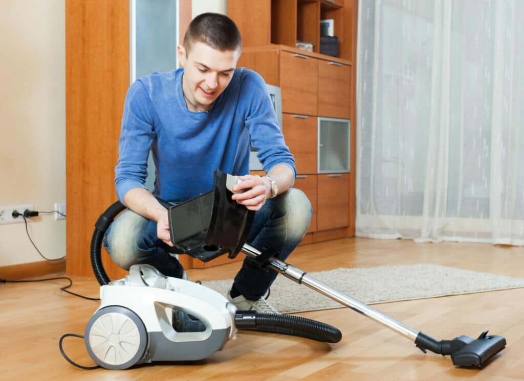A man crouching while cleaning a vacuum cleaner filter, with the vacuum and nozzle on a hardwood floor.