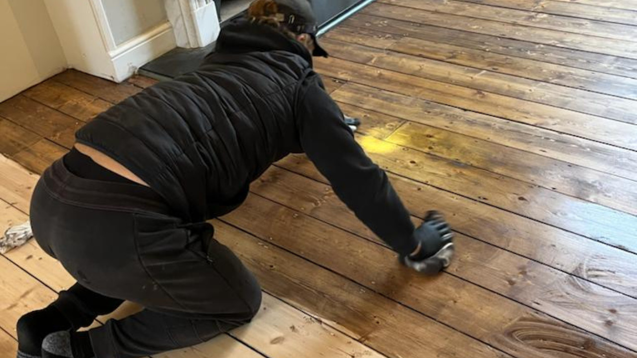 A professional flooring specialist hand-applying dark wood floor staining to Victorian pine softwood floorboards to ensure deep penetration.