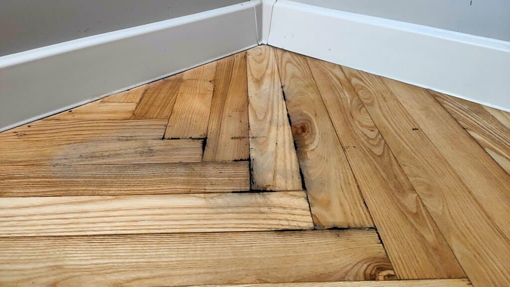 Close-up of a corner section of a parquet floor showing signs of water damage, with dark stains and slight warping of the wood. The image emphasizes the need for professional parquet flooring restoration to address such issues. This image is featured in the Mr Sander® blog post focused on parquet flooring restoration.