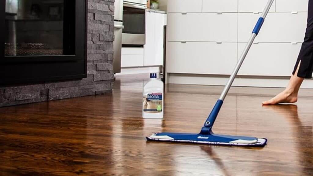 A freshly refinished dark hardwood floor being cleaned with a mop and Bona hardwood floor cleaner. The floor reflects light, highlighting its smooth and polished finish. A modern kitchen and stone fireplace are visible in the background, showcasing a contemporary and elegant interior design.