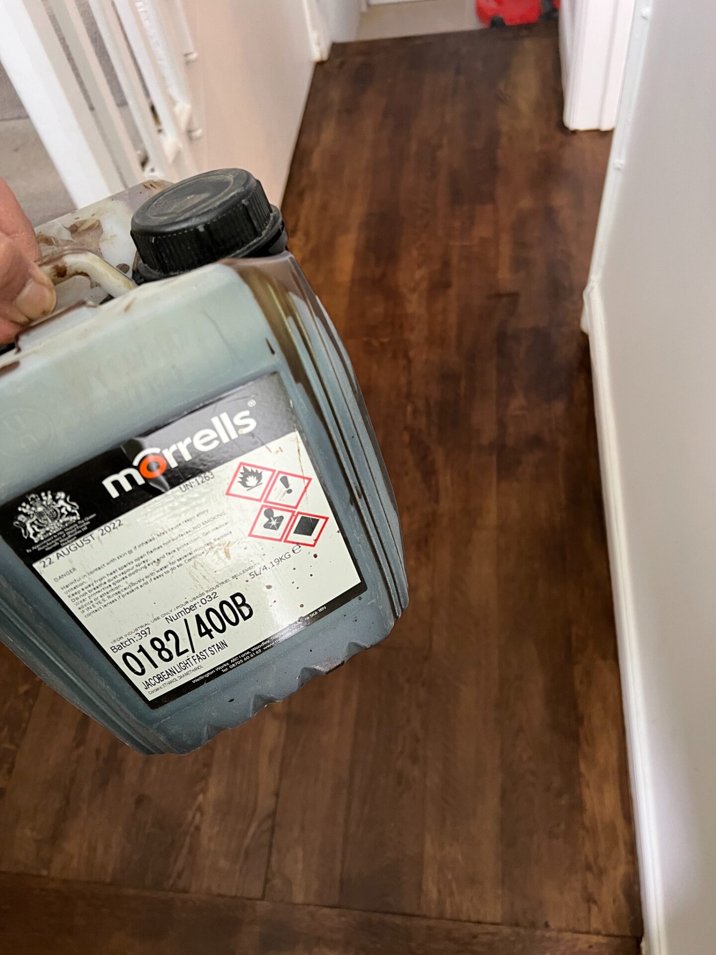 Close‑up of a tradesperson holding a Morrells wood‑stain can above freshly stained dark‑walnut hallway floorboards.