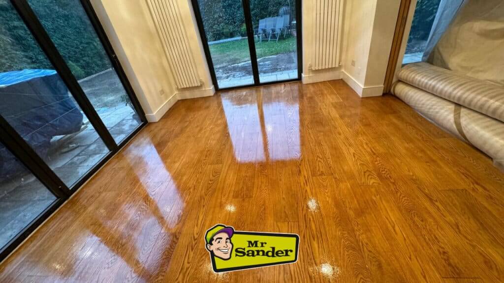 A beautifully refinished oak hardwood floor with a glossy finish, showcasing its rich color and natural wood grain. The Mr Sander® logo is featured prominently.