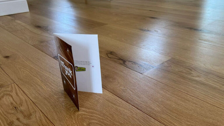 Close-up of a newly finished oak floor with a “Thank you” card standing upright; tight joints, light reflecting from the boards, smooth satin sheen.