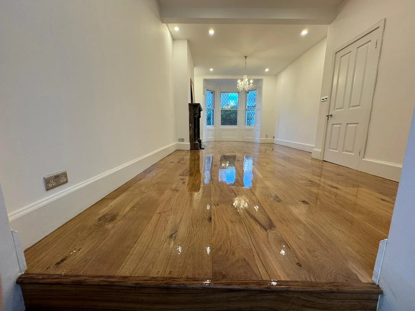Open-plan living room with newly refinished oak hardwood flooring, high-gloss lacquer reflecting bay window and chandelier.
