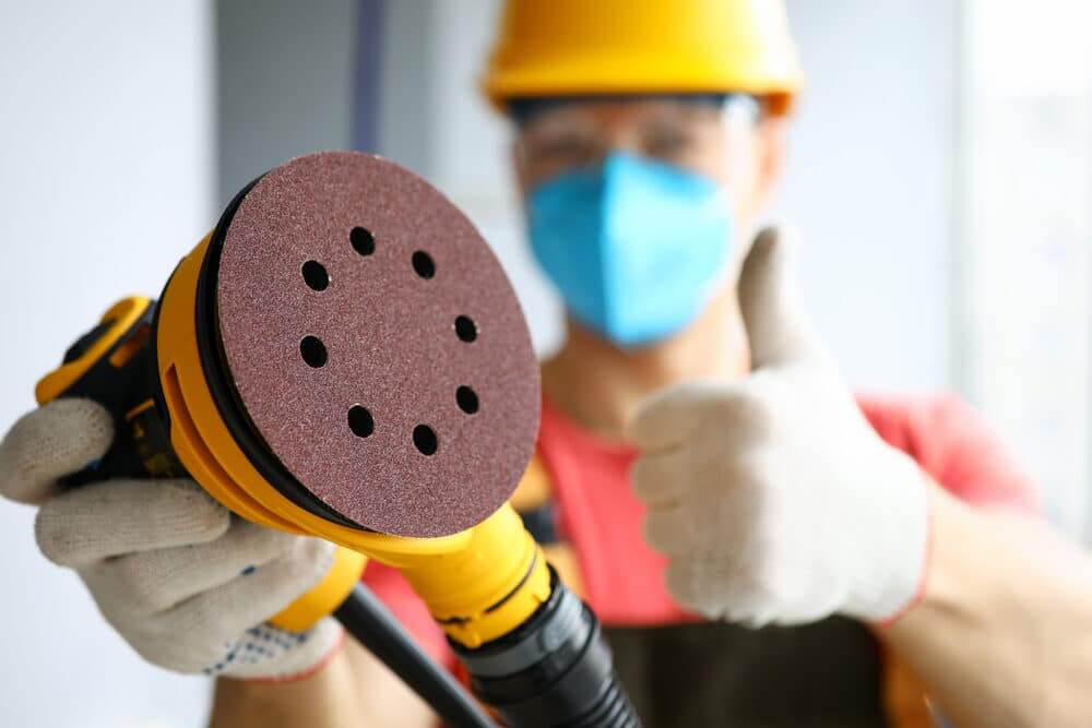 A professional in safety gear, including a hard hat, mask, and gloves, holding a yellow orbital sander with a close-up of the sanding pad. The worker gives a thumbs-up, ready for floor sanding and refinishing, ensuring a high-quality result for oak hardwood floors.