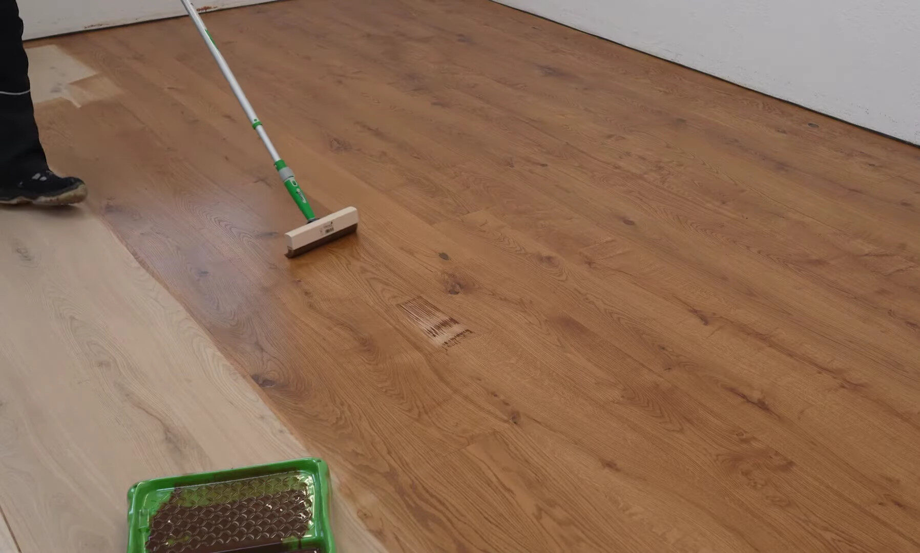 A person applies an oil-based finish to a wooden floor using a long-handled applicator. The floor shows a clear contrast between the sanded, untreated section on the left and the glossy, finished section on the right. A green tray with the oil-based finish, likely Osmo oil, is visible in the foreground.