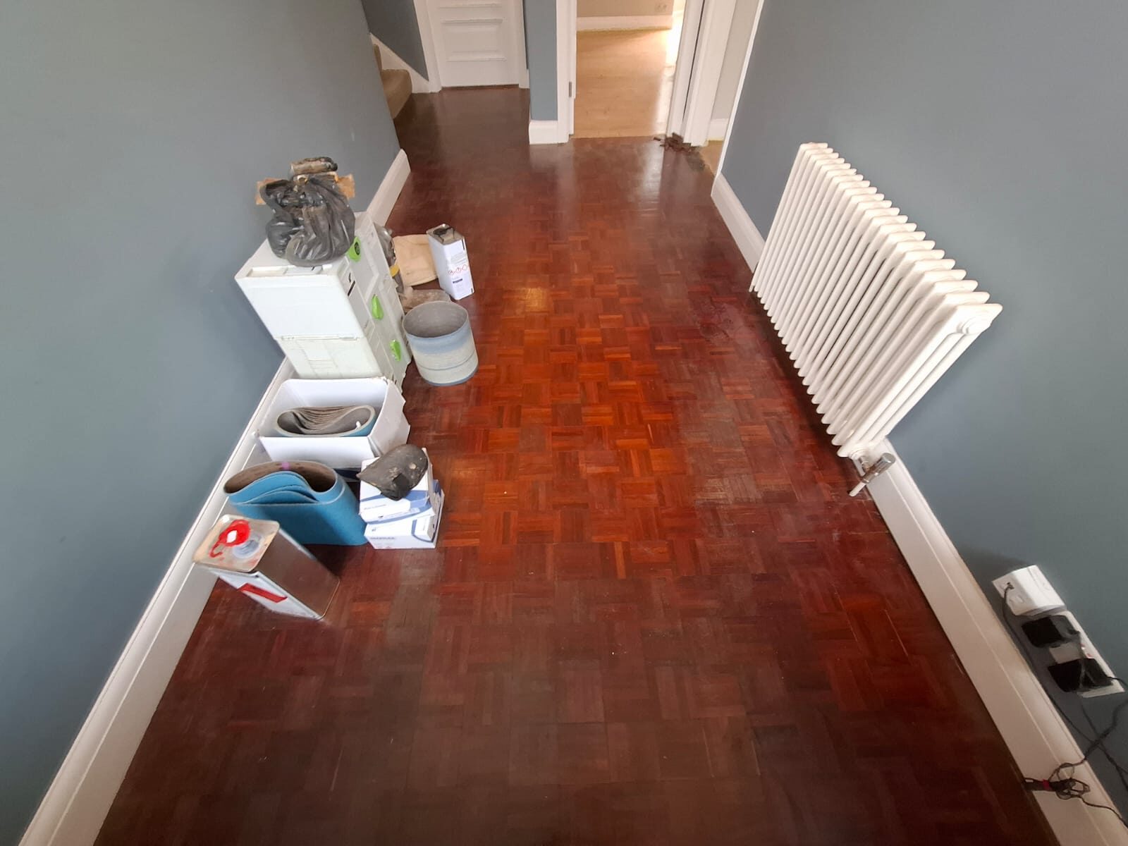 Parquet Floor Refinishing prep stage: worn mosaic parquet hallway with sanding equipment and materials set out before restoration.